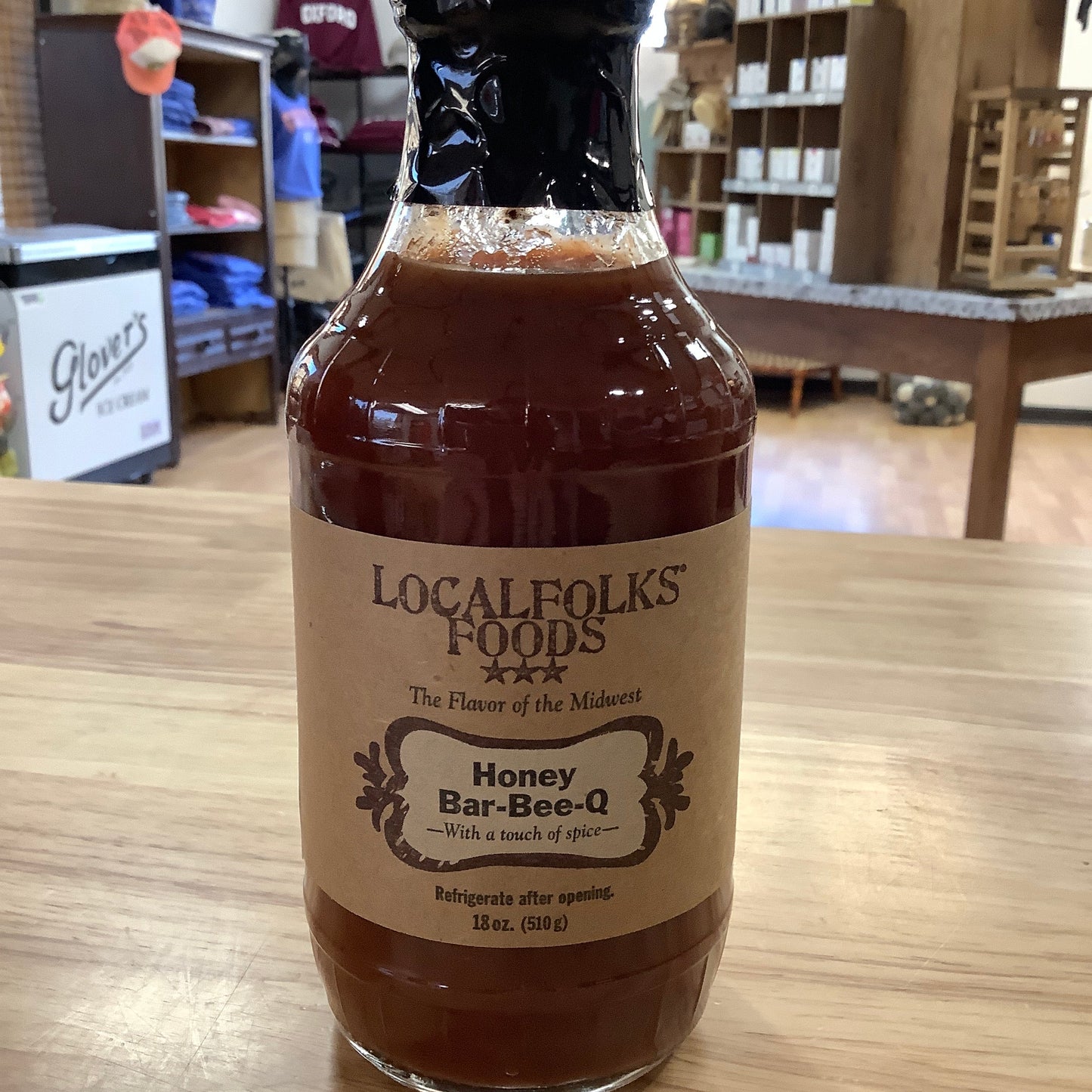 Bottle of Localfolks Foods Honey Bar-B-Q sauce on a wooden surface with a blurred indoor background.